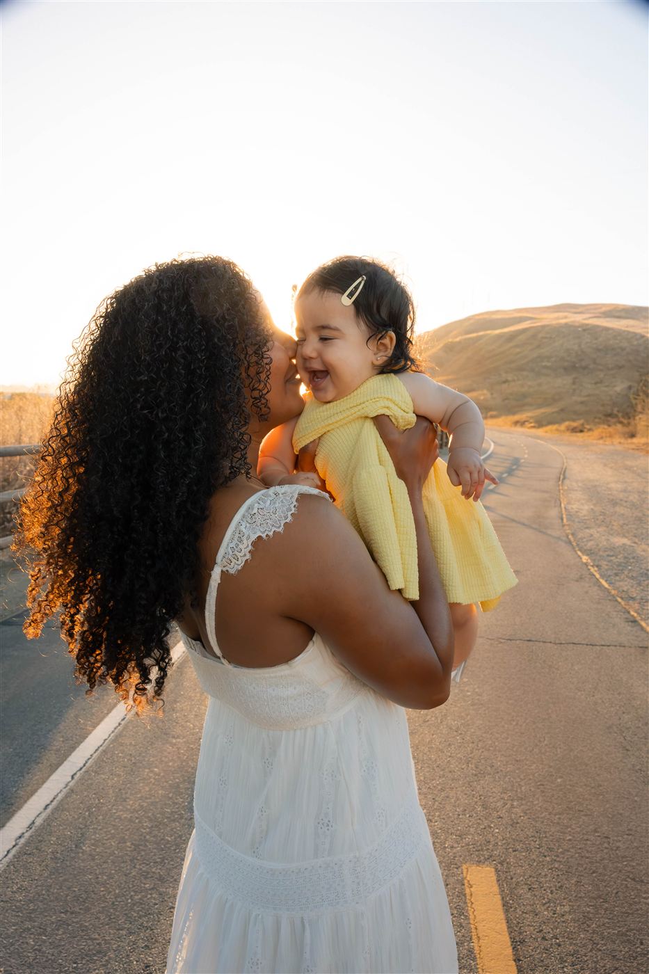 Mother in a white lace dress kissing her laughing baby daughter on a golden hillside road at sunset