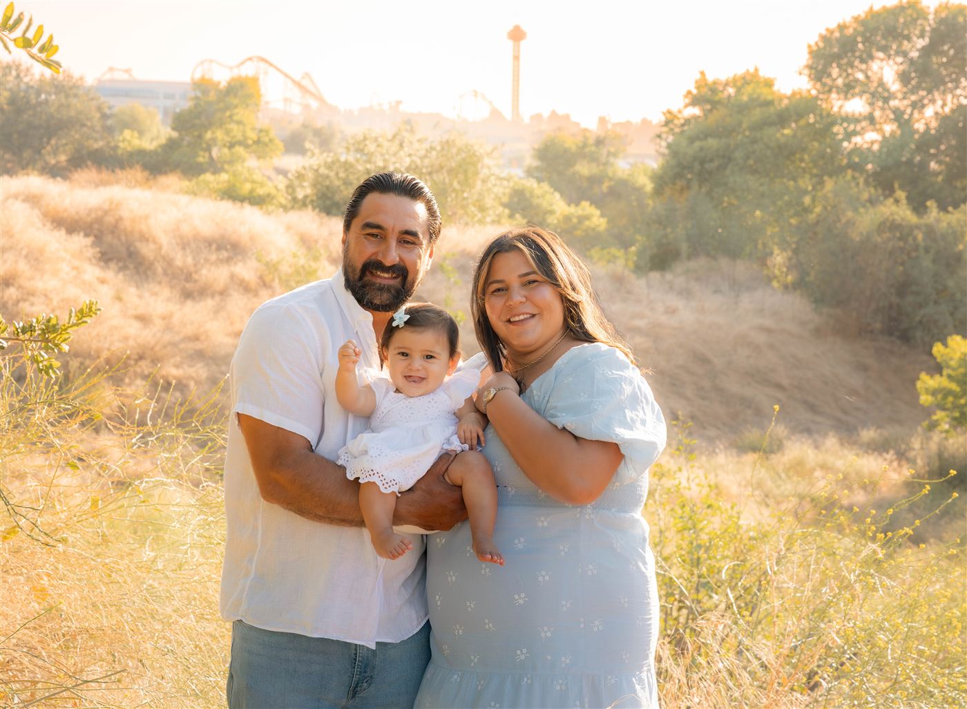 The DeAlba family smiling and holding their baby daughter in a golden hillside meadow at sunset